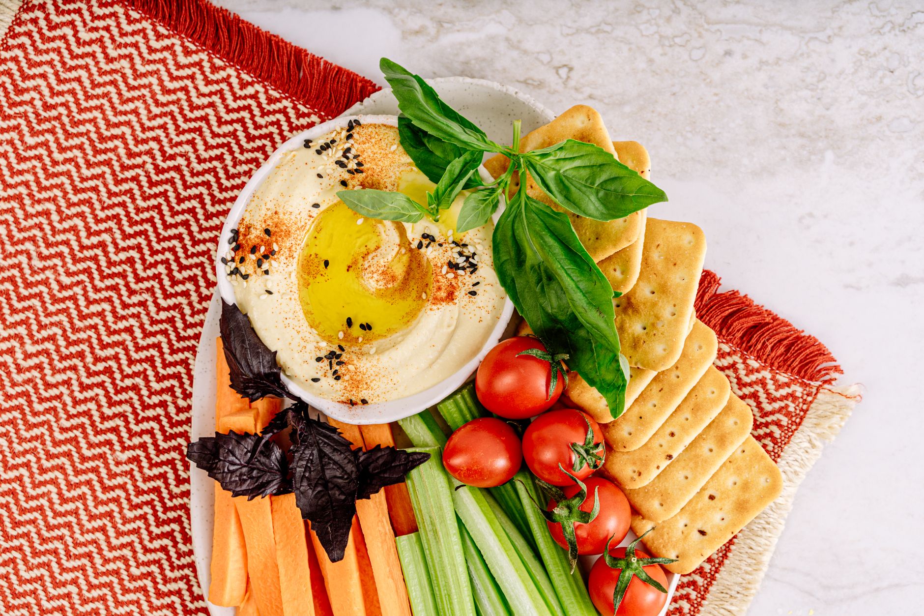 Top-down view of hummus with crackers, fresh vegetables, and herbs on a colorful platter.