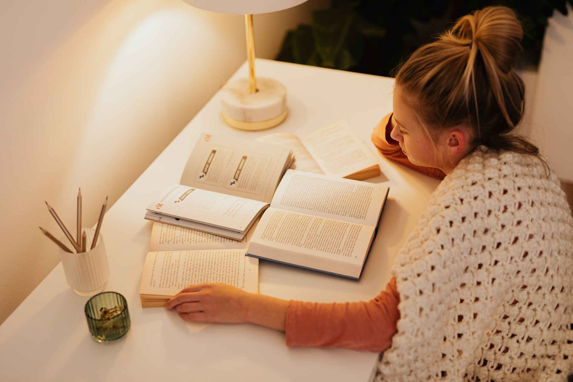 A woman enjoys a quiet evening reading multiple books at a warmly lit desk.