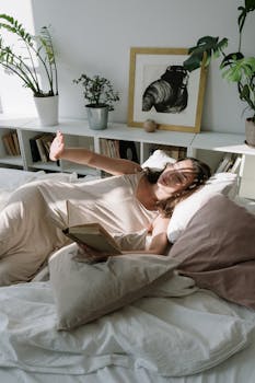 Asian woman enjoying a tranquil morning reading in bed with soft sunlight.