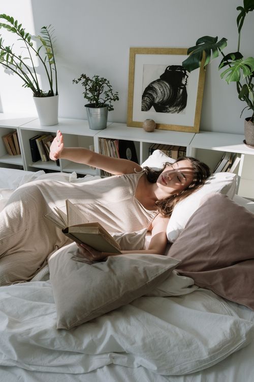 Free Asian woman enjoying a tranquil morning reading in bed with soft sunlight. Stock Photo