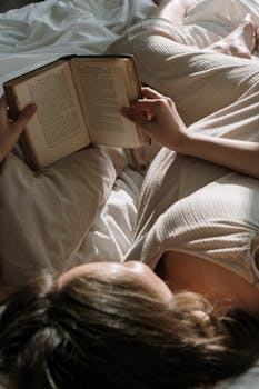 A woman lying in bed reading a book in soft, natural light, creating a peaceful atmosphere.