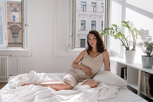 Free Woman with Hand on Belly Sitting on a Bed with White Linen Stock Photo
