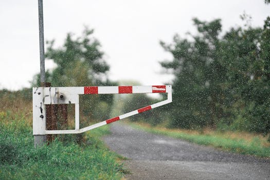 Old white barrier gate on a rural road surrounded by lush greenery, featuring a dynamic summer rain scene.