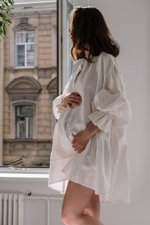 Free Pregnant Woman in White Dress Near a Window Stock Photo