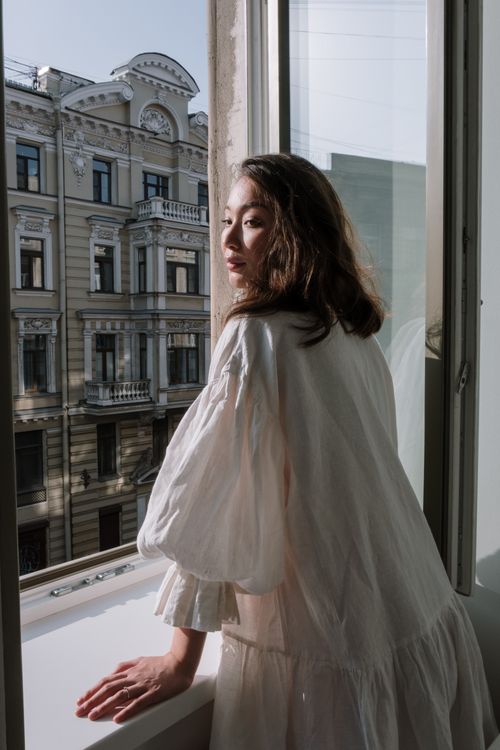 Free Portrait of a woman in a white dress gazing out from an open window. Stock Photo