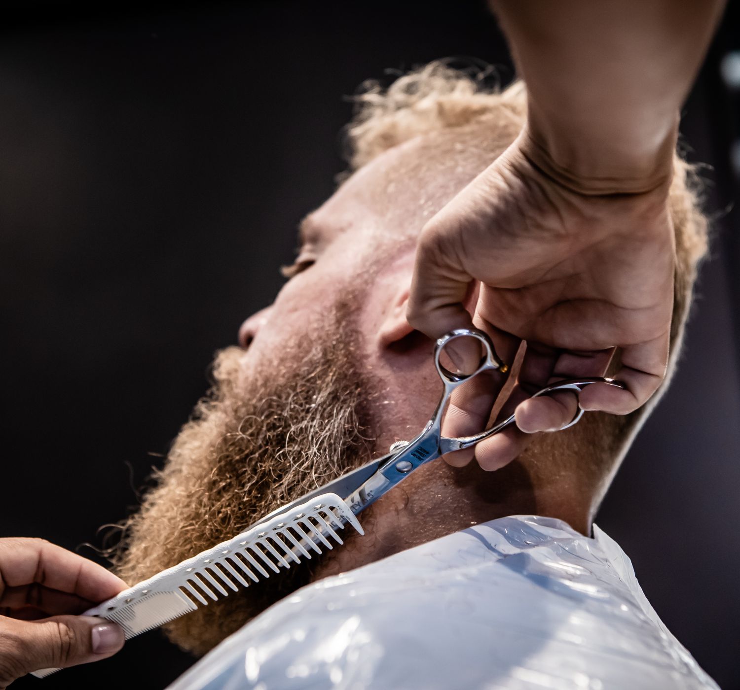 Close-up of a barber trimming a client's beard with scissors and comb.
