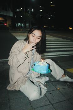 A young woman sits on a city street with a birthday cake at night.