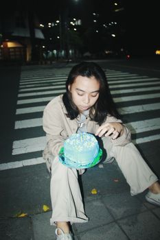 A young woman sits on the sidewalk holding a birthday cake at night in the city.