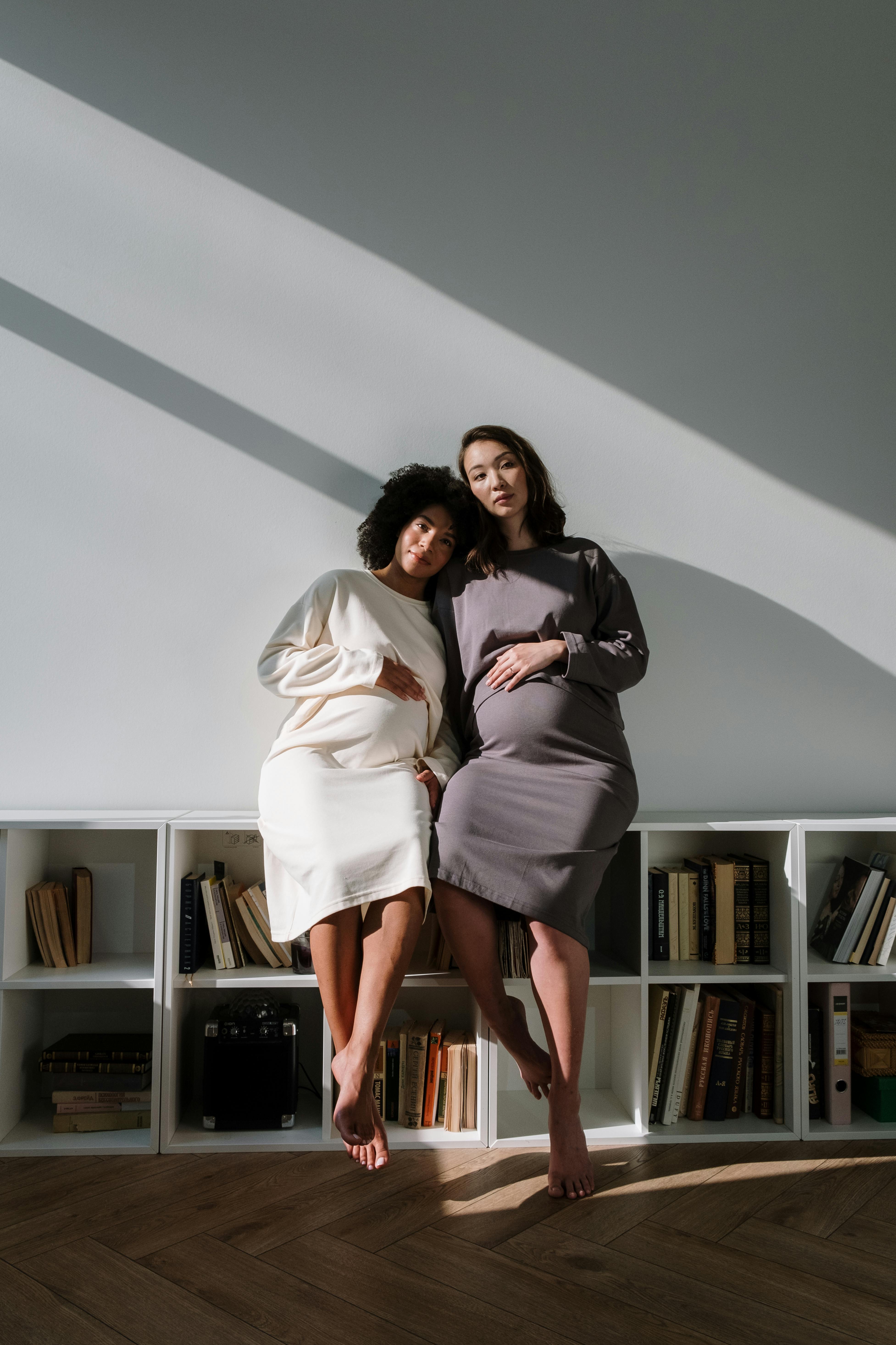 2 pregnant women in midi dresses sitting on bookshelf