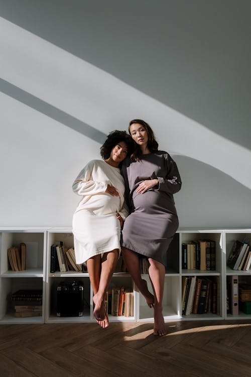 2 Pregnant Women in Midi Dresses Sitting on Bookshelf