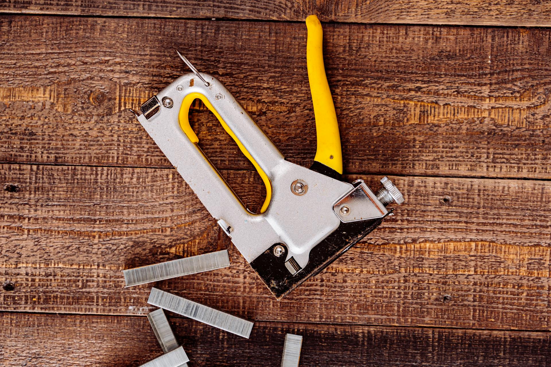 A staple gun with yellow handle and staples on a rustic wooden surface, captured from above.