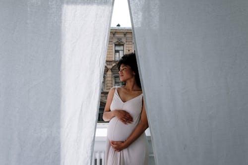 Woman Standing Behind the White Curtain Holding Her Baby Bump while Looking Afar