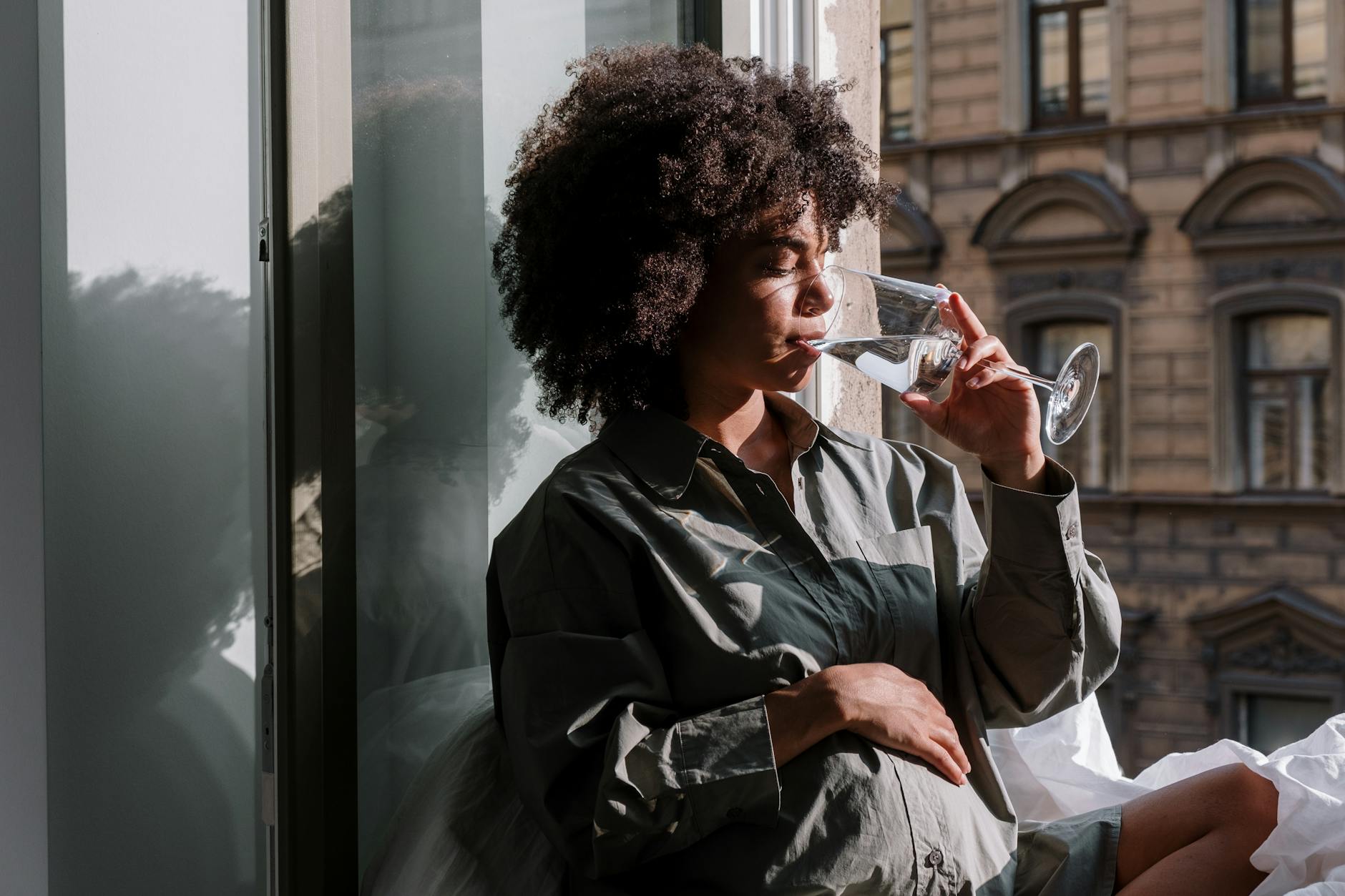 Pregnant woman in long sleeves enjoying a glass of water by a window indoors.