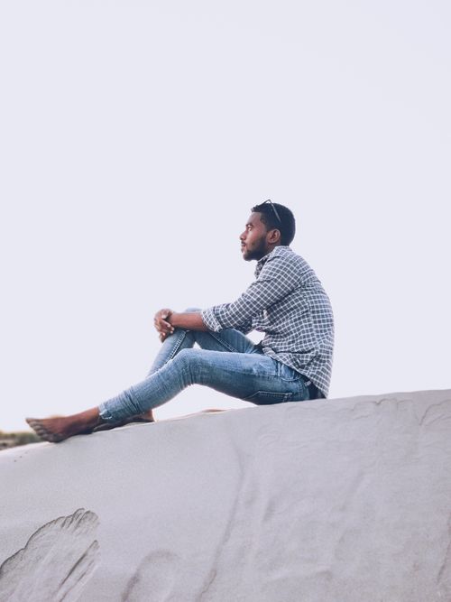 Free Side view of a man sitting on a sand dune, gazing afar against a clear sky. Stock Photo