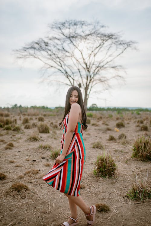 Free Woman in Colorful Dress Standing on a Drought Field Near Leafless Tree while Posing at the Camera Stock Photo