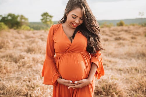 Woman in Orange Long Sleeve Dress Looking at the Baby Bump she is Holding while Standing on Brown Grass Field