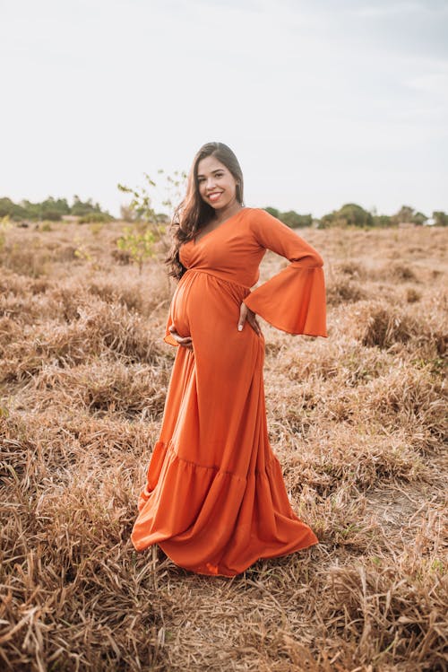 Woman in Orange Long Sleeves Dress Standing on Brown Grass Field while Smiling at the Camera