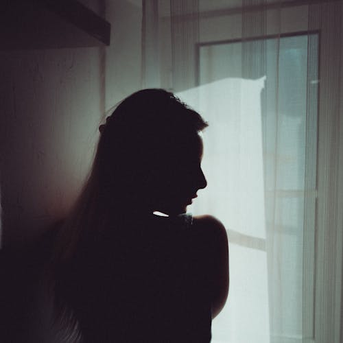 Back view of anonymous female touching hair while standing against window with bright sunlight
