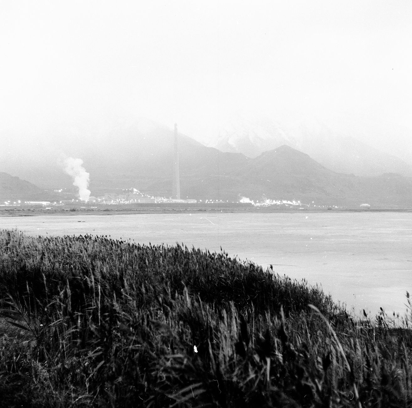 Black and white landscape featuring industrial plant against mountain backdrop near water.
