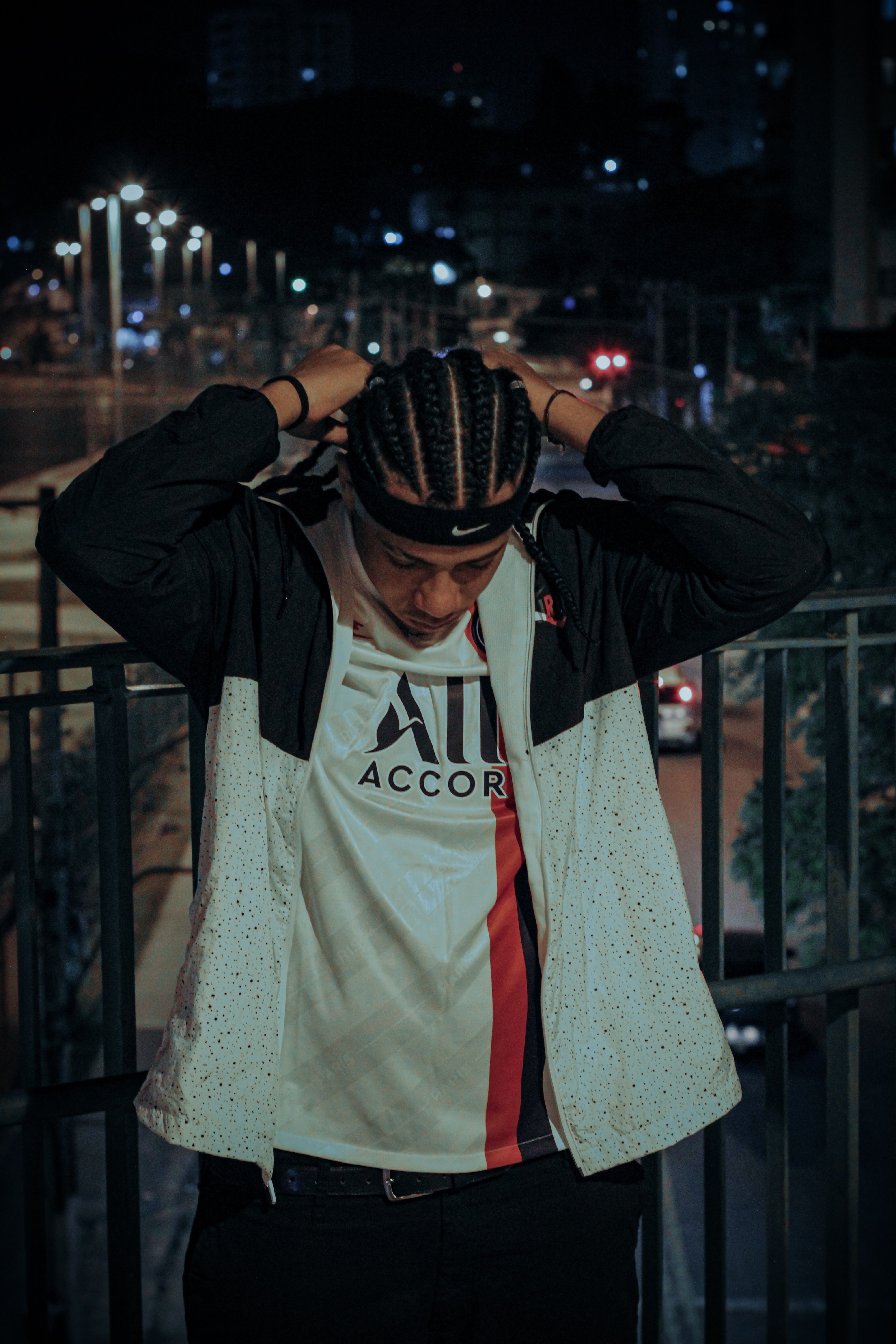 A young man with braided hair standing near metal railings during nighttime in the city.