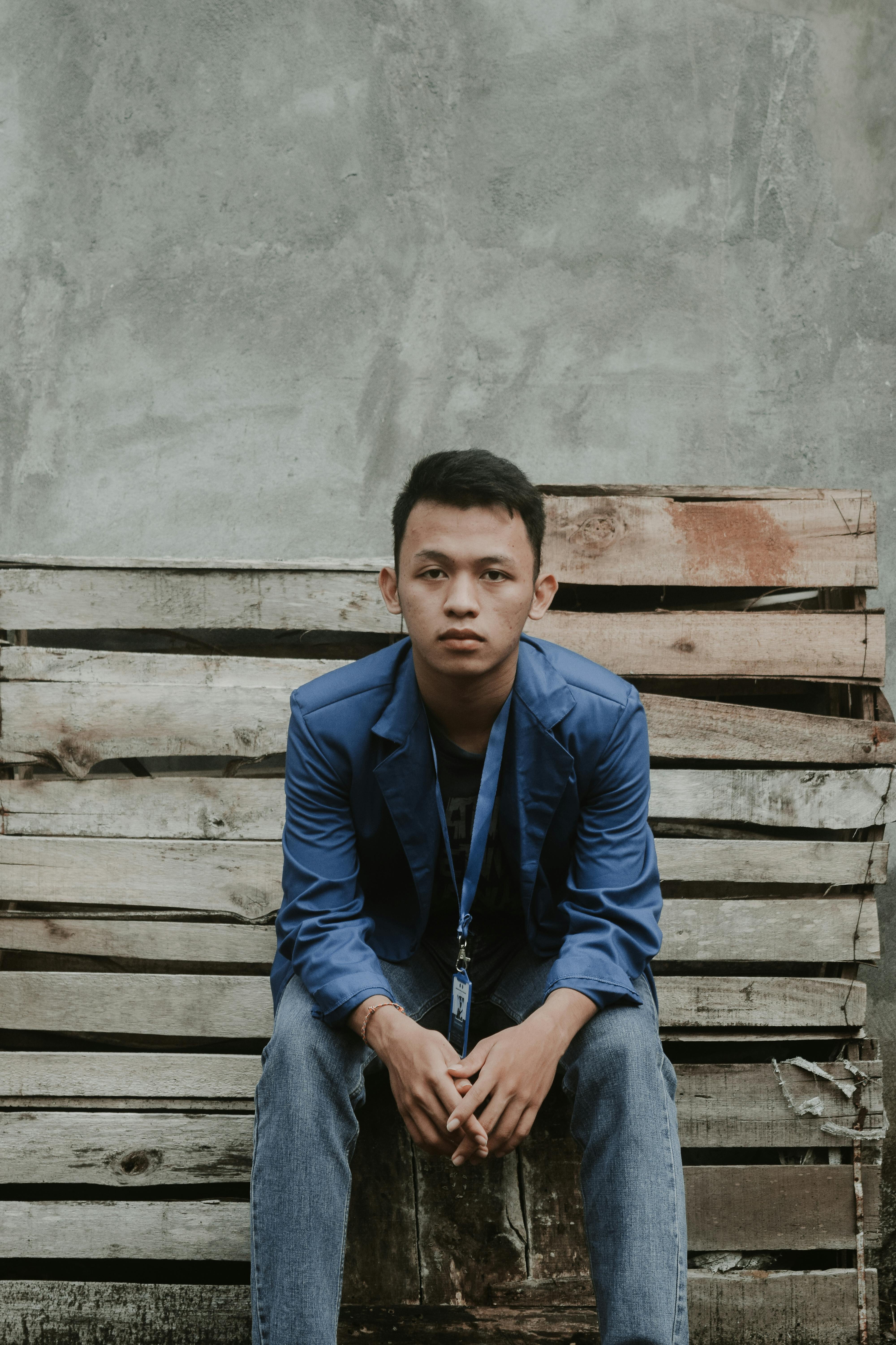 concentrated young ethnic guy sitting on weathered bench and looking at camera near concrete wall