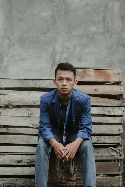 Concentrated young ethnic guy sitting on weathered bench and looking at camera near concrete wall