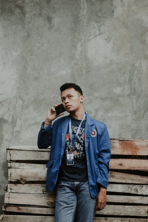 Thoughtful young Asian guy with dark hair in casual clothes and name tag leaning on shabby wooden bench an talking on smartphone on street