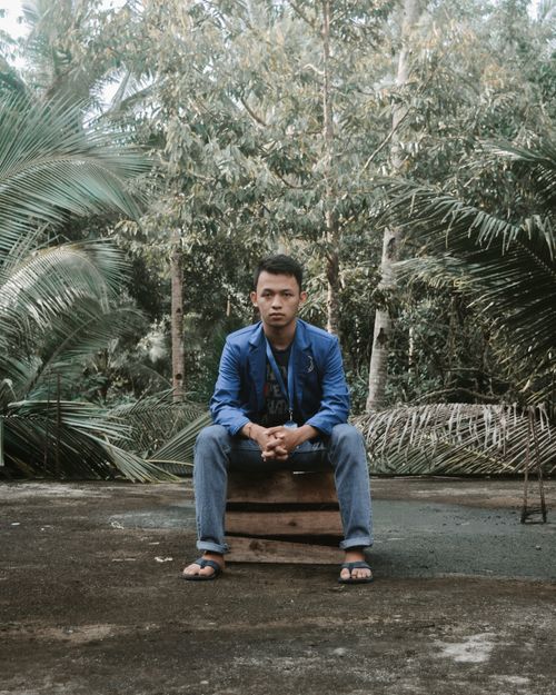 Free Full body of serious young Asian male model in casual clothes sitting on pallet box and looking at camera against lush topical trees in rainforest Stock Photo