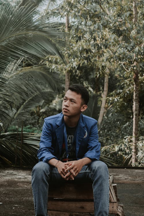 Pensive Asian male teenager with dark hair in casual clothes sitting on pallet box and looking away pensively against lush green tropical trees in forest
