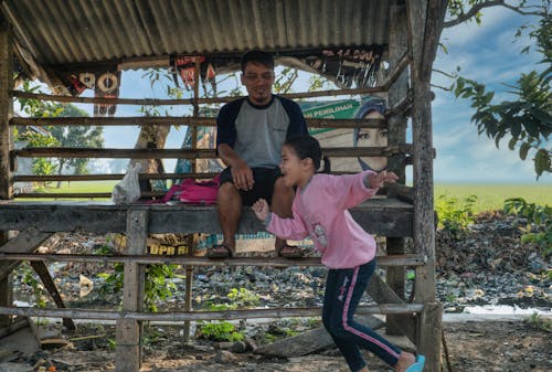 Free Side view of cheerful little Asian girl in casual clothed smiling and running while spending time with happy father sitting in rustic wooden cabin in village Stock Photo