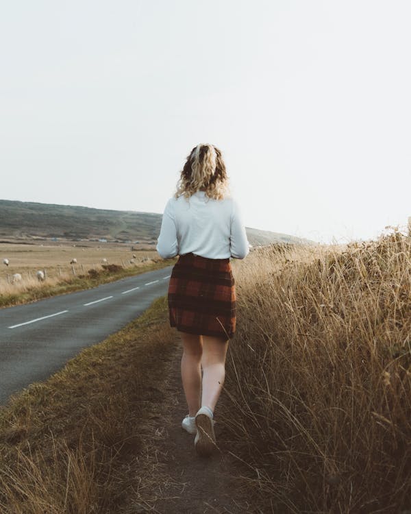 Free Back view of unrecognizable female in checkered skirt walking on empty asphalt road in hilly terrain Stock Photo