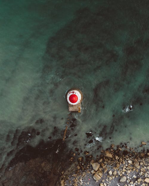 Top view of lighthouse with red tower located in calm azure water near stony shore in seaside
