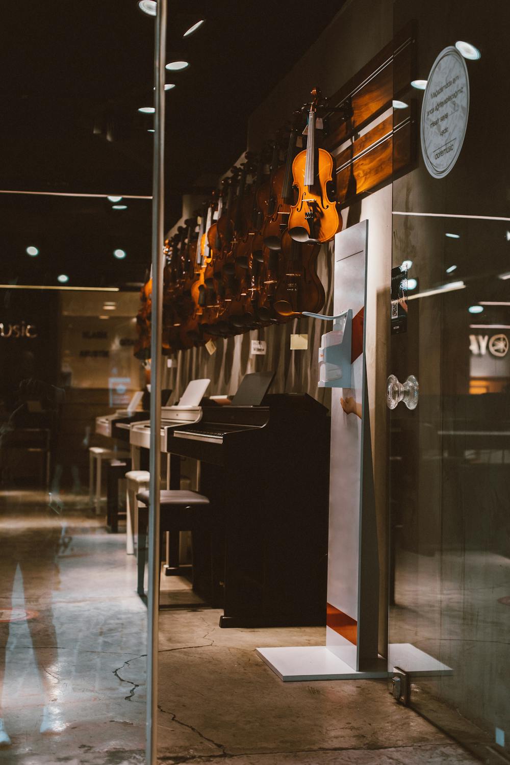 Glass door open to a music store with a row of small upright pianos and violins hanging above.