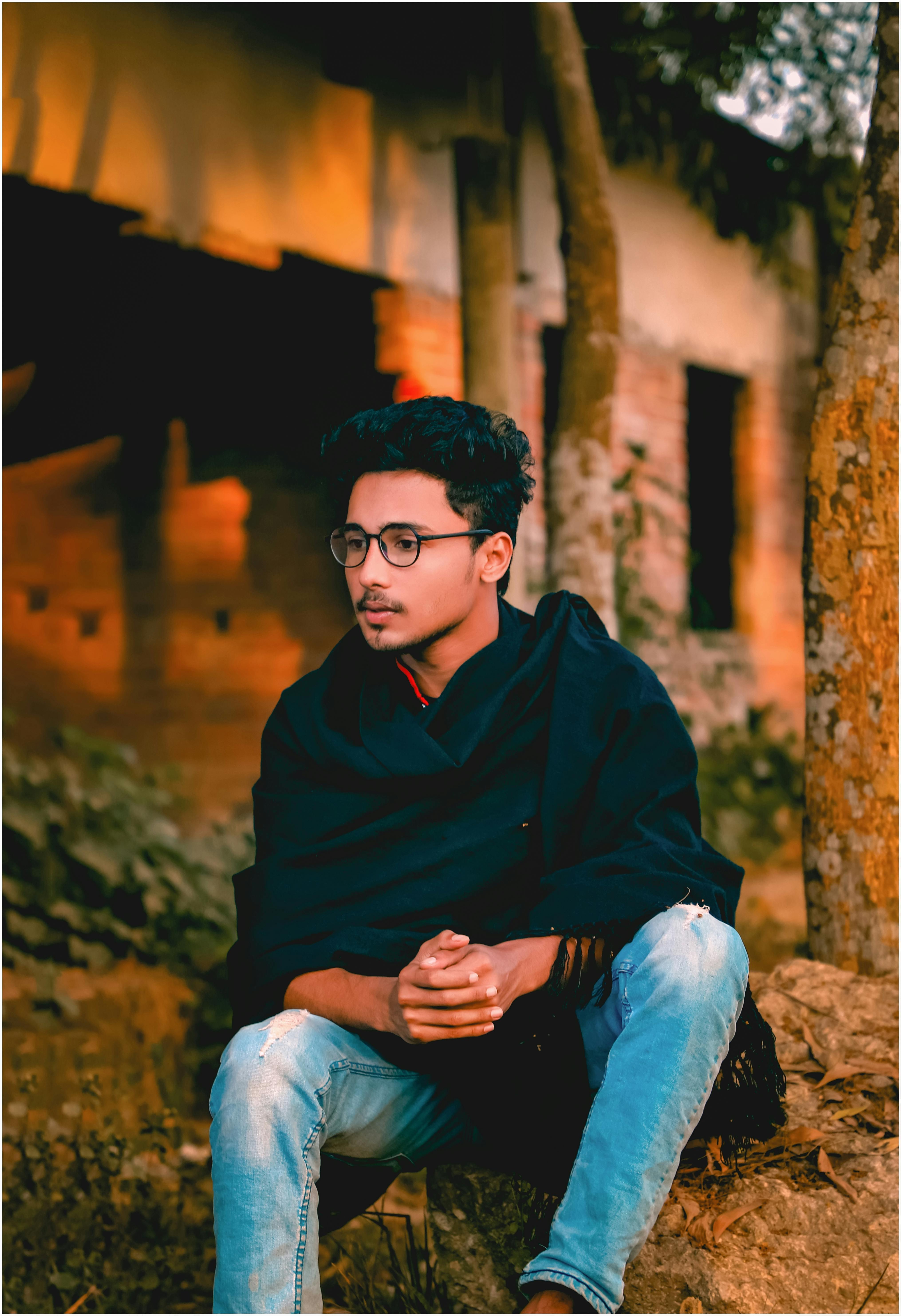 Young man with glasses sitting on a rock, wearing jeans in a rustic setting.