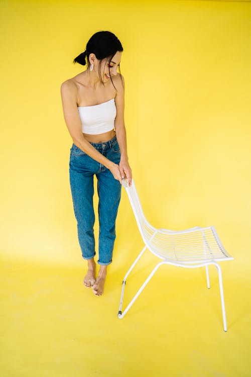 Mujer En Camiseta Blanca Sin Mangas Y Pantalones Vaqueros Azules Sosteniendo Silla De Plástico Blanco