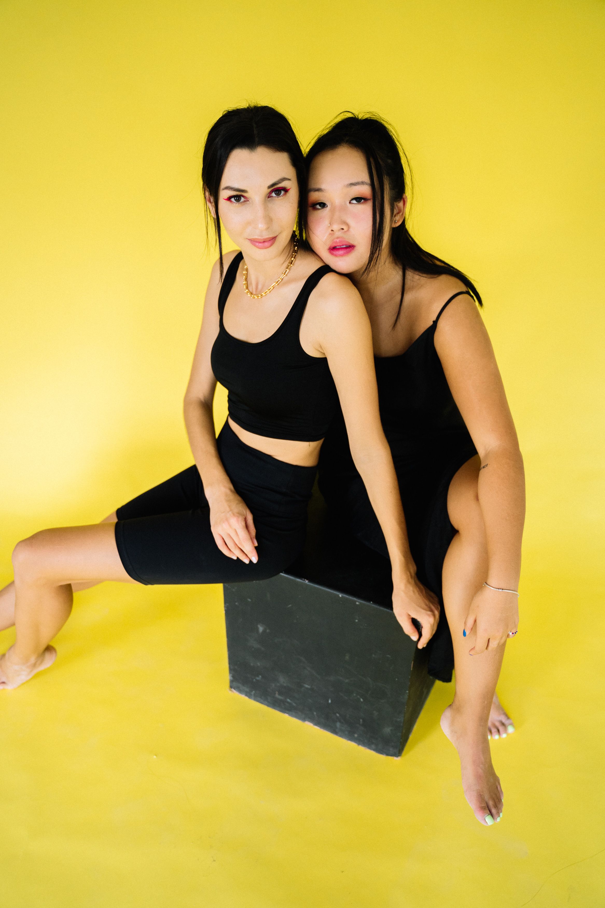 两名身穿黑衣的女性坐在一个黑匣子上，背景是亮黄色