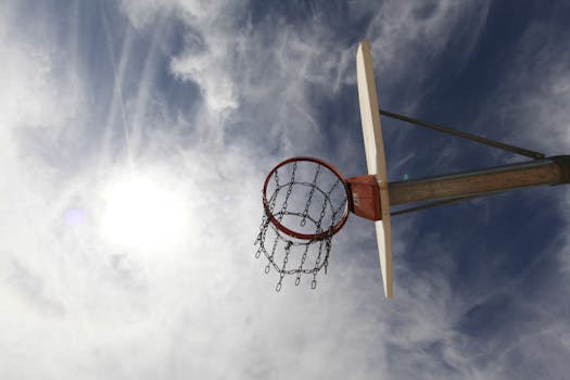 A basketball hoop with a chain net is silhouetted against a cloudy blue sky, creating a dynamic sports scene.