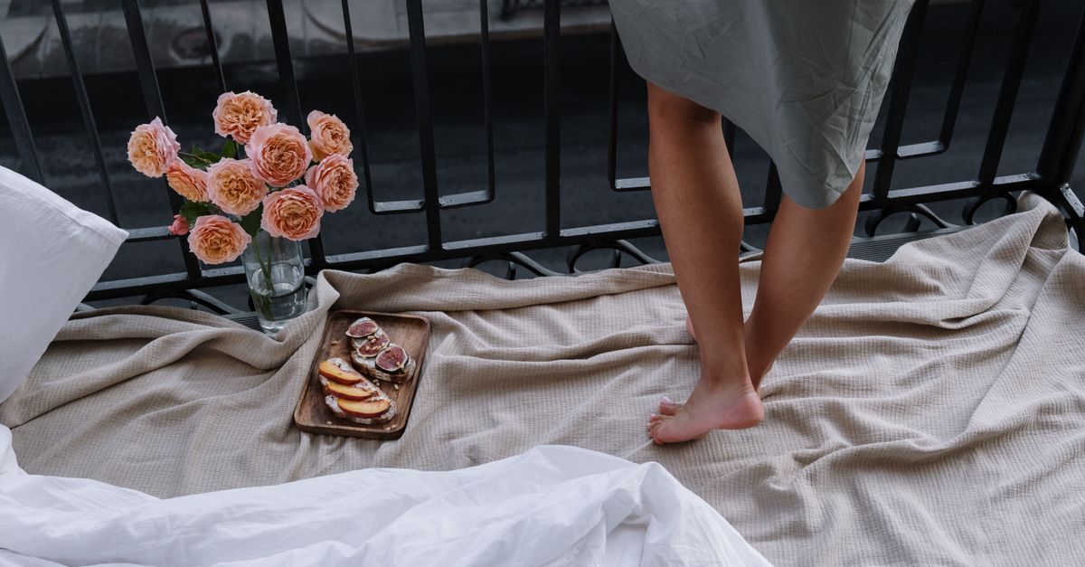 A woman stands barefoot on a balcony with a cozy setting, flowers, and breakfast.