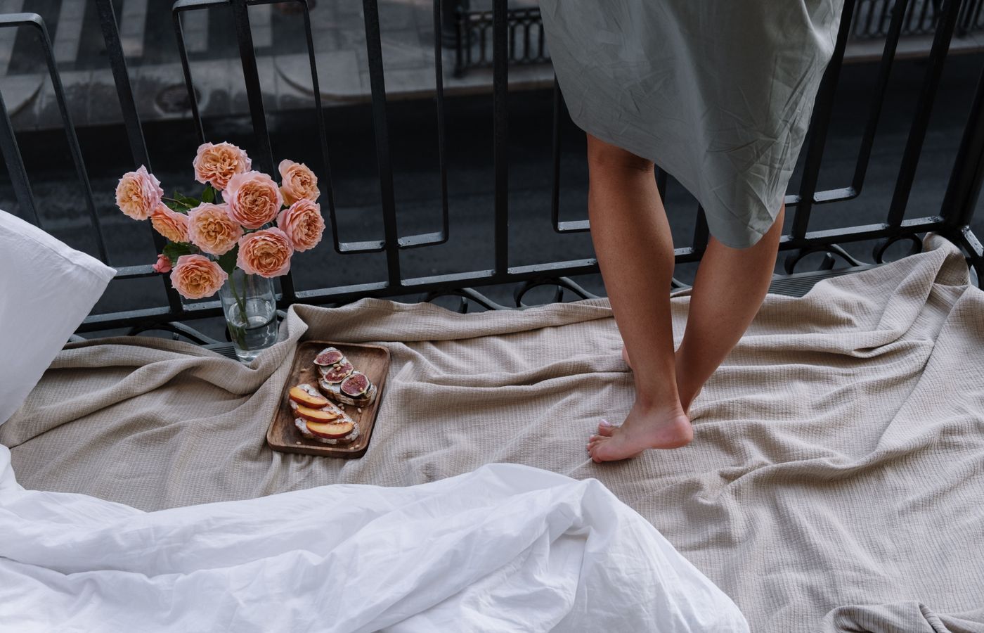 A woman stands barefoot on a balcony with a cozy setting, flowers, and breakfast.