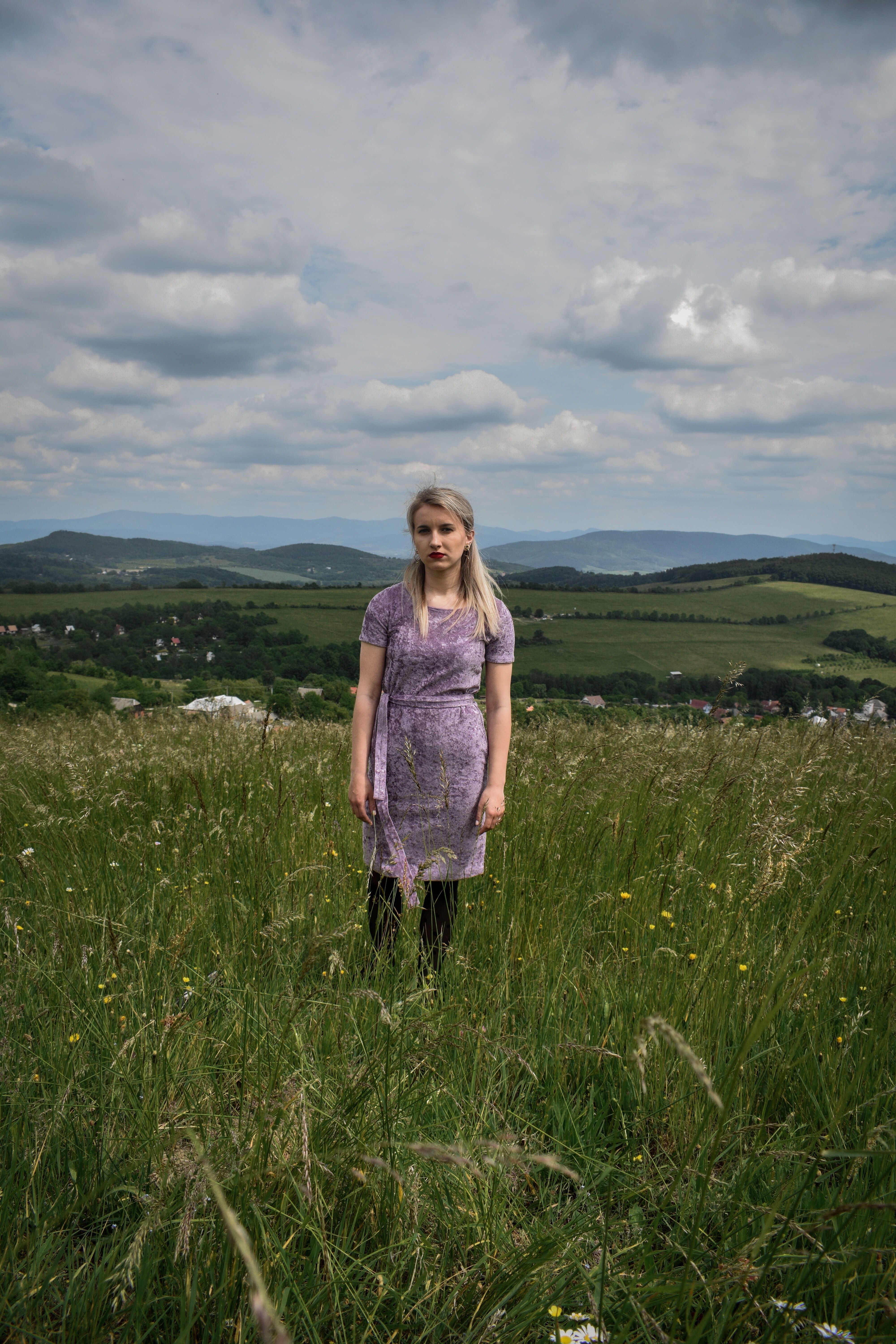 A serene outdoor portrait of a woman in a purple dress standing in a picturesque field under a cloudy sky.