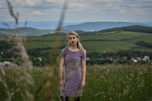 A woman in a purple dress stands in a rural field with a mountain backdrop.
