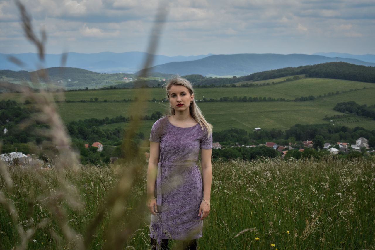a woman in a purple dress stands in a rural field with a mountain back