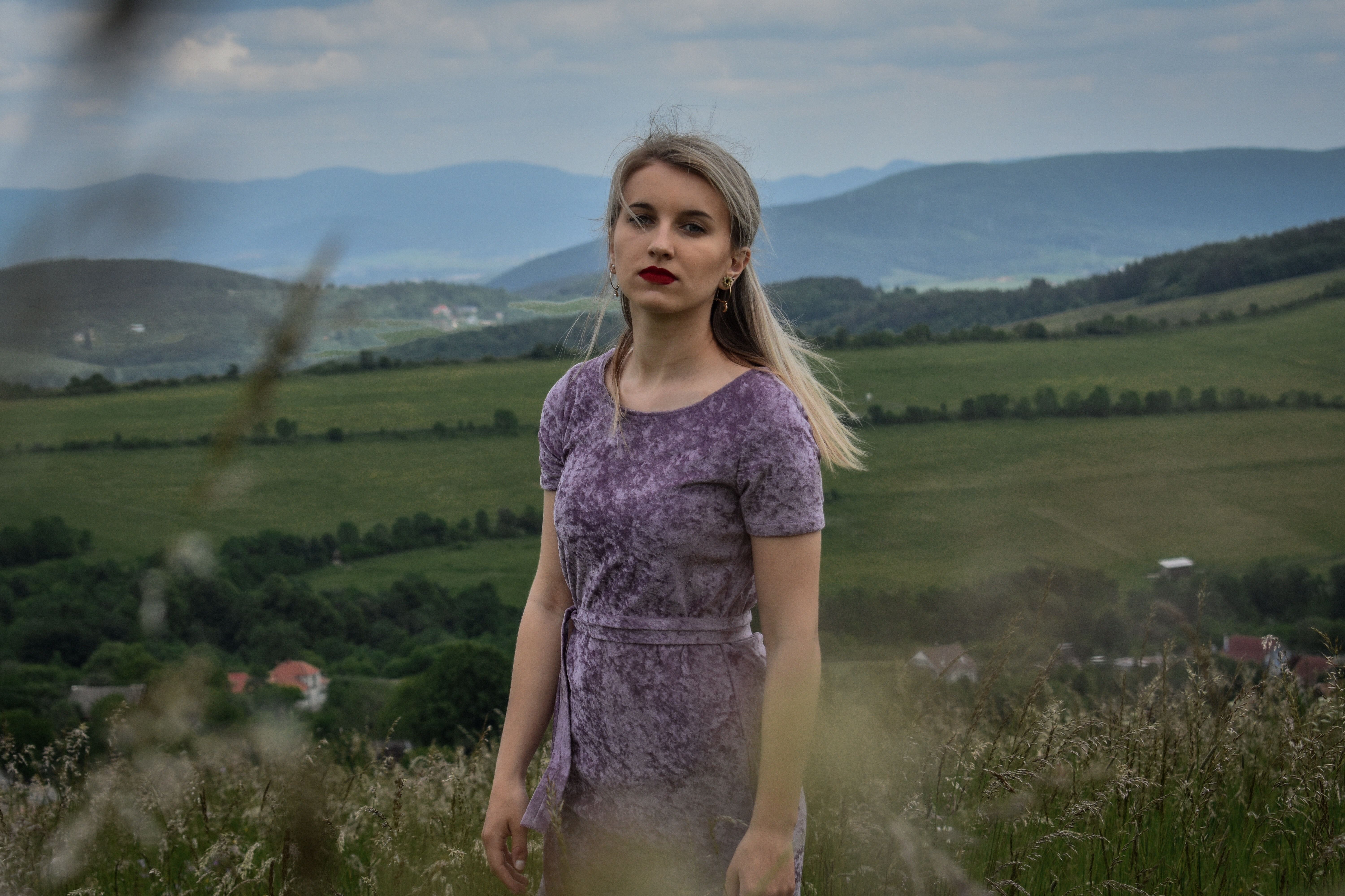 Portrait of a woman in a lilac dress standing in a scenic meadow with mountains in the background.