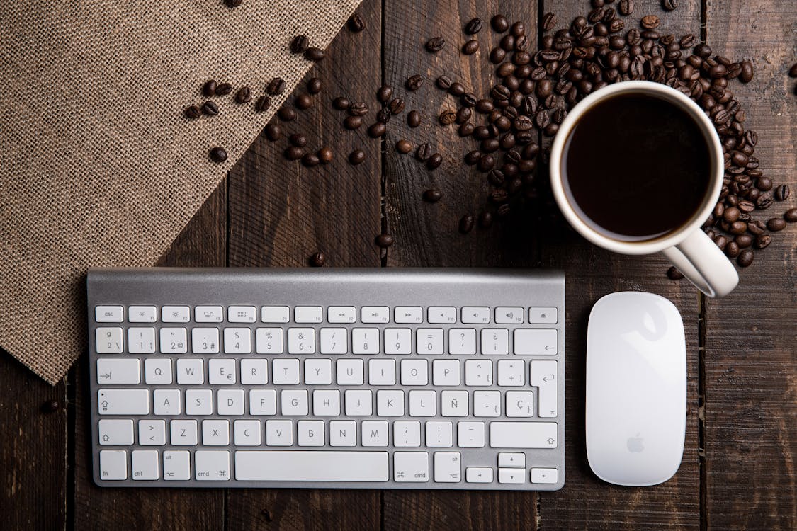豆の横にコーヒーで満たされたアップルマジックキーボード、マウス、マグカップのフラットレイ写真