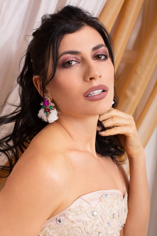 Side view of young female with stylish makeup and earrings wearing dress touching hair and looking at camera