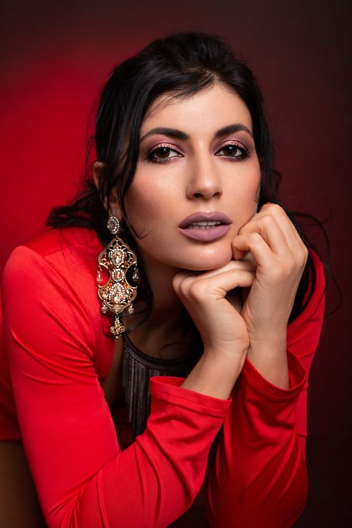 Free Confident female with makeup and dark hair wearing red outfit and earrings with necklace leaning on hand while looking at camera in studio Stock Photo