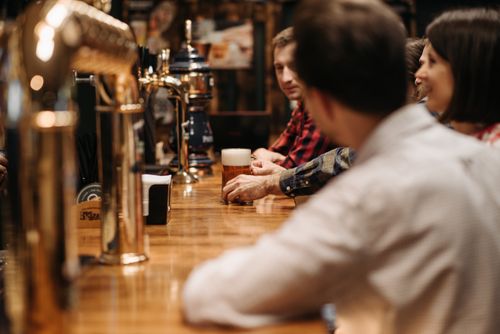 Free Friends enjoying a beer at a bustling bar with wooden counter and brass taps. Stock Photo