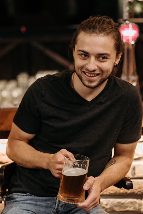 Free A man in a black shirt smiles while holding a mug of beer in an indoor bar setting. Stock Photo