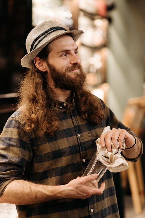 Free A bearded bartender with long hair and a hat cleaning a glass in a cozy indoor setting. Stock Photo