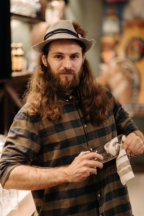 Free A bearded bartender in checkered shirt and hat, cleaning a glassware in a rustic pub setting. Stock Photo